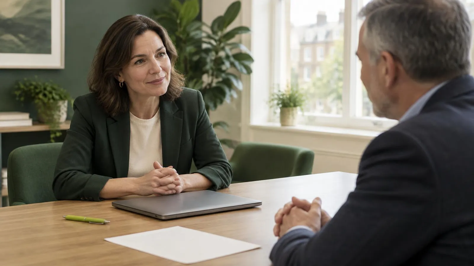 A founder in her forties and a prospective client in his fifties talking across a meeting table in a bright UK office, a closed laptop between them and a single sheet of paper face down, a lime-green pen on the table, soft morning light.